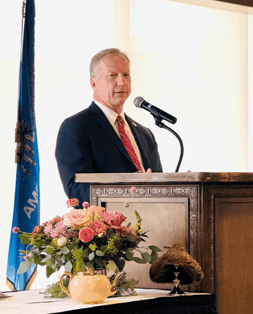 Kevin Hern speaking at event, wearing formal suit and red tie, standing behind podium with flowers.